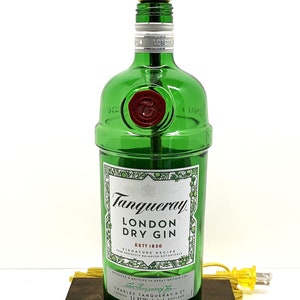 May include: A green glass bottle lamp with a gold metal lampshade. The bottle is labeled "Tanqueray London Dry Gin Est. 1830". The lamp is sitting on a dark brown wooden base.