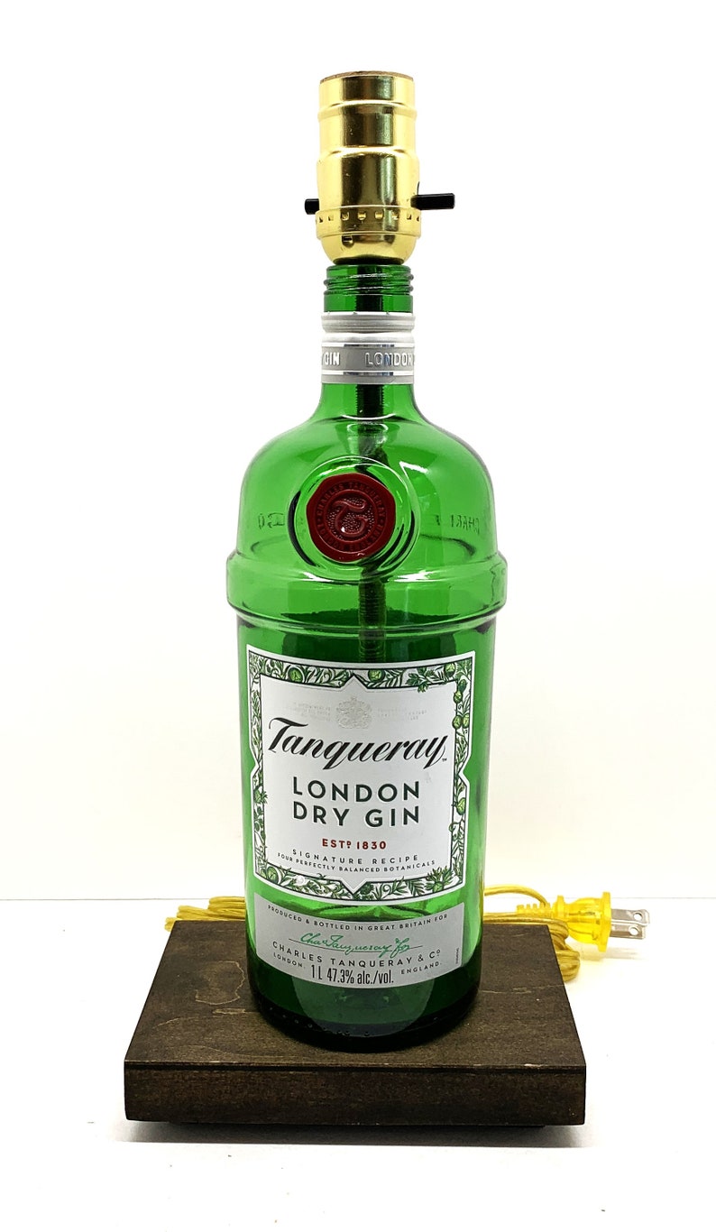 May include: A green glass bottle lamp with a gold metal lampshade. The bottle is labeled "Tanqueray London Dry Gin Est. 1830". The lamp is sitting on a dark brown wooden base.