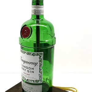 May include: A green glass bottle lamp with a gold-colored lampshade and a black switch. The bottle is labeled "Tanqueray London Dry Gin Est. 1830". The lamp is sitting on a dark brown wooden base and has a yellow electrical cord with a yellow plug.