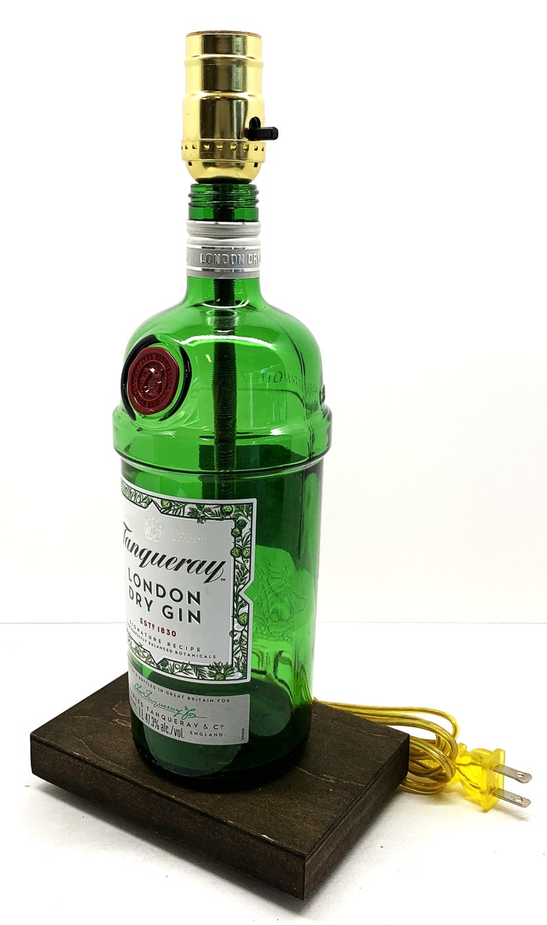 May include: A green glass bottle lamp with a gold-colored lampshade and a black switch. The bottle is labeled "Tanqueray London Dry Gin Est. 1830". The lamp is sitting on a dark brown wooden base and has a yellow electrical cord with a yellow plug.