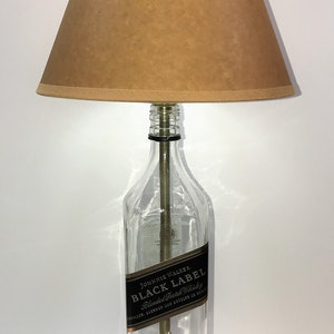 May include: A table lamp made from a repurposed Johnnie Walker Black Label bottle with a brown lampshade. The bottle is clear glass with a black label that reads "JOHNNIE WALKER. BLACK LABEL Blended Scotch Whisky DISTILLED, BLENDED AND BOTTLED IN SCOTLAND AGED 12 YEARS". The lamp has a wooden base.