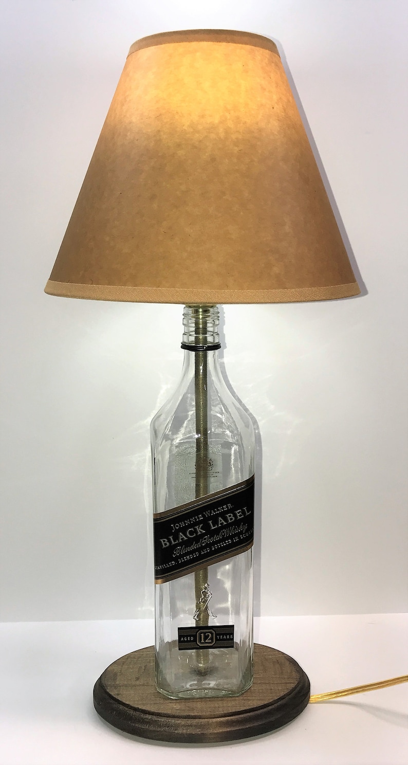 May include: A table lamp made from a repurposed Johnnie Walker Black Label bottle with a brown lampshade. The bottle is clear glass with a black label that reads "JOHNNIE WALKER. BLACK LABEL Blended Scotch Whisky DISTILLED, BLENDED AND BOTTLED IN SCOTLAND AGED 12 YEARS". The lamp has a wooden base.
