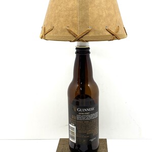May include: A unique lamp crafted from a Guinness Extra Stout beer bottle. It features a tan lampshade with brown leather-like accents and a wooden base. The lamp includes a brown power cord and plug.