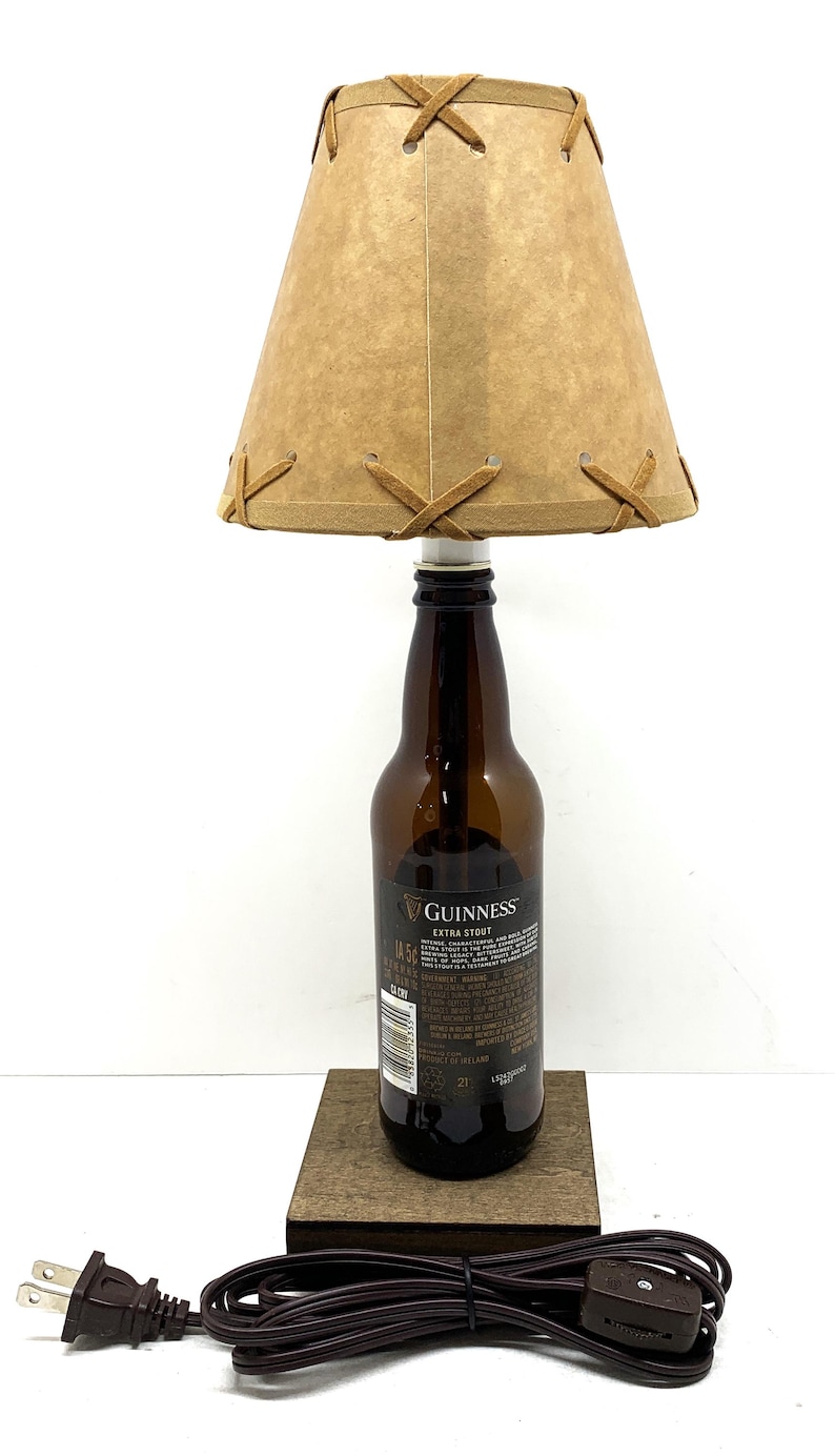 May include: A unique lamp crafted from a Guinness Extra Stout beer bottle. It features a tan lampshade with brown leather-like accents and a wooden base. The lamp includes a brown power cord and plug.