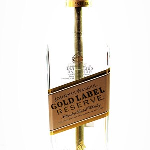 May include: A clear glass bottle lamp with a gold label that reads "Johnnie Walker Gold Label Reserve Blended Scotch Whisky". The bottle is sitting on a wooden base. The lamp has a gold light bulb socket and a yellow cord.