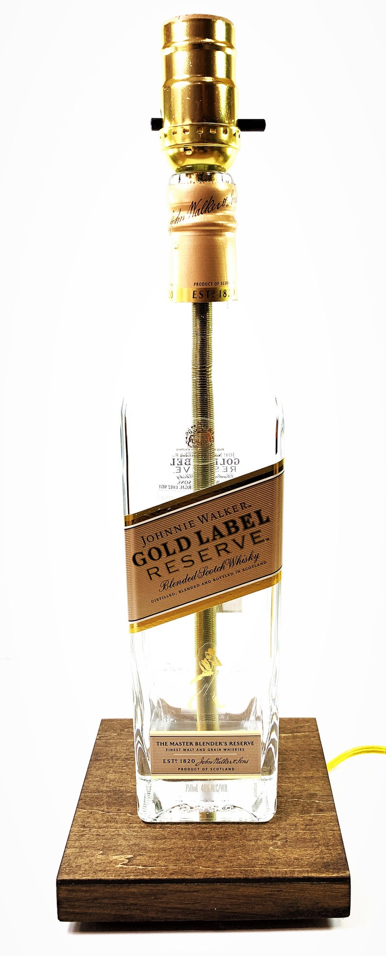 May include: A clear glass bottle lamp with a gold label that reads "Johnnie Walker Gold Label Reserve Blended Scotch Whisky". The bottle is sitting on a wooden base. The lamp has a gold light bulb socket and a yellow cord.