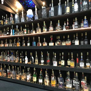 May include: A collection of various liquor bottles displayed on multiple shelves. The bottles are of different shapes and sizes, with a variety of labels and colors. The shelves are dark, and the bottles are illuminated by overhead lighting.
