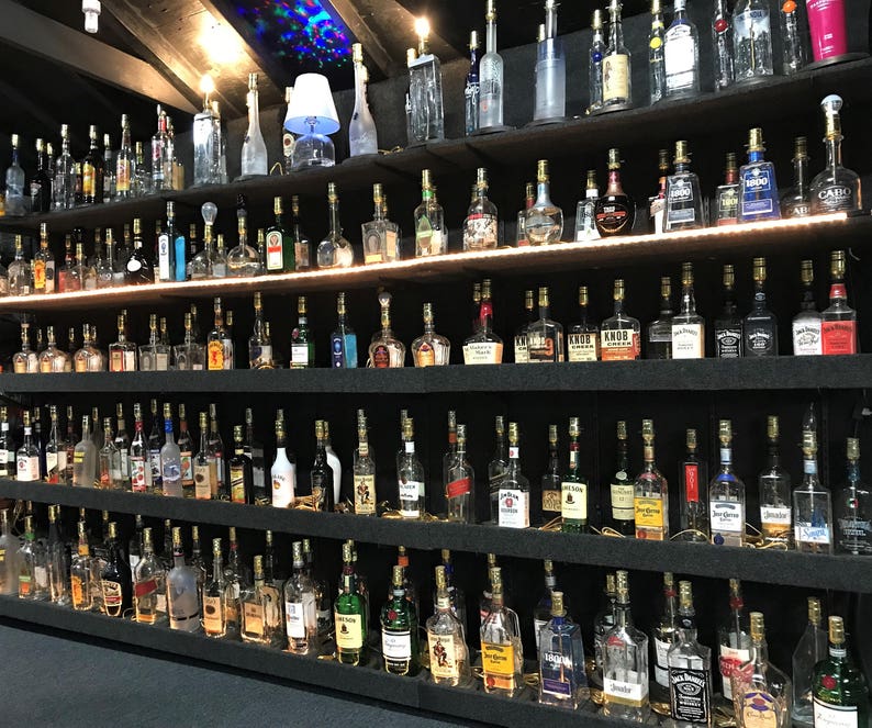 May include: A collection of various liquor bottles displayed on multiple shelves. The bottles are of different shapes and sizes, with a variety of labels and colors. The shelves are dark, and the bottles are illuminated by overhead lighting.