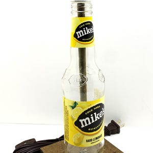 May include: A repurposed lamp made from a Mike's Hard Lemonade bottle. The bottle features a yellow and black label with lemon graphics. It has a white lamp shade and sits on a small wooden base. A brown cord and plug are attached.