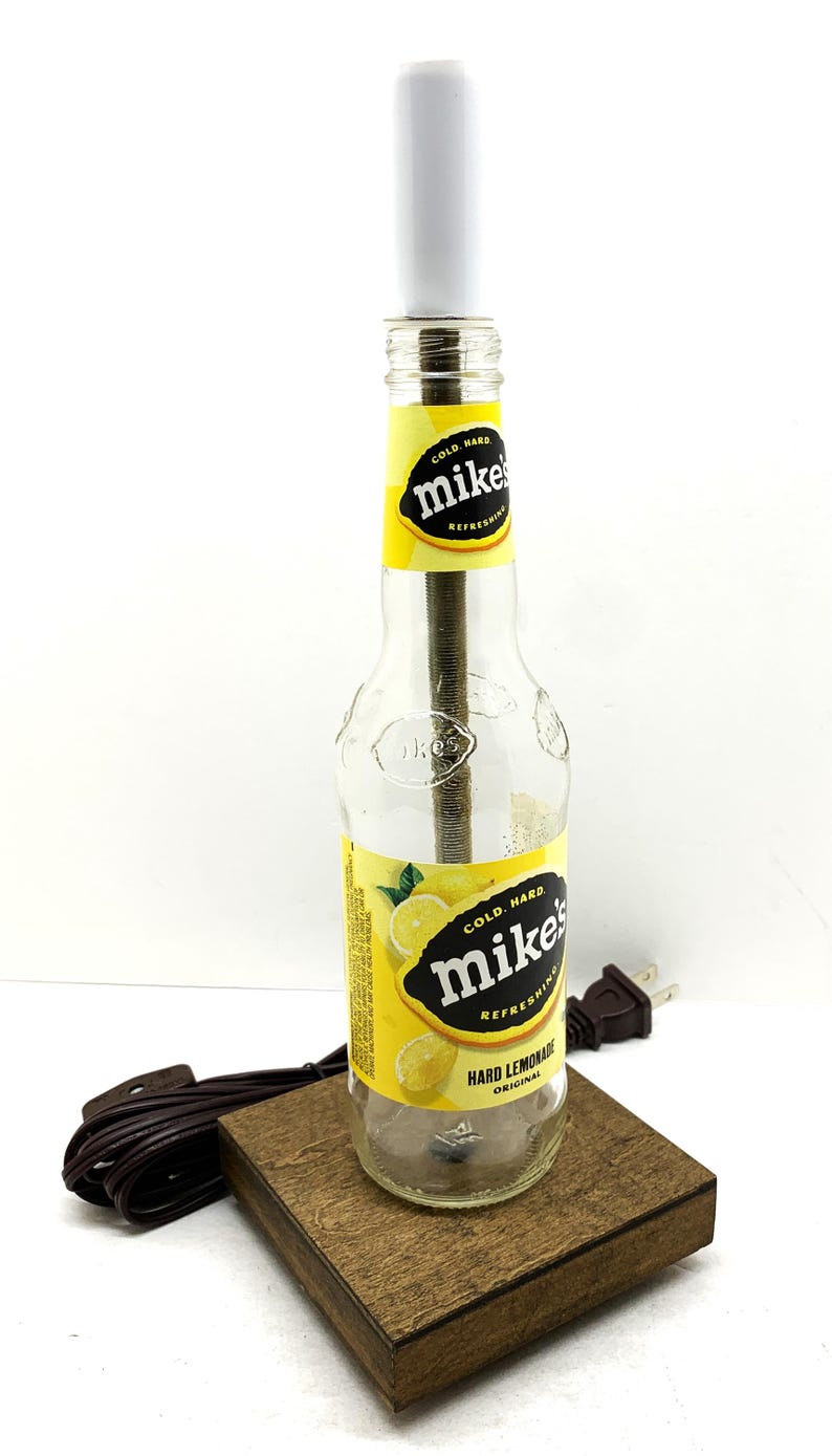 May include: A repurposed lamp made from a Mike's Hard Lemonade bottle. The bottle features a yellow and black label with lemon graphics. It has a white lamp shade and sits on a small wooden base. A brown cord and plug are attached.