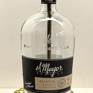 May include: A unique lamp made from a repurposed tequila bottle. The clear glass bottle features black labels with the "el Mayor" logo and "Blanco Tequila" text. The lamp has a gold-colored bulb socket and sits on a dark wooden base.