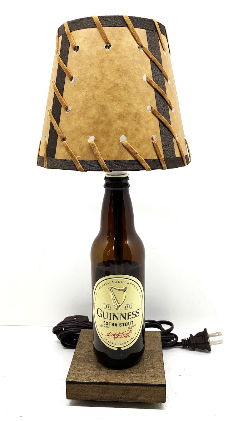 May include: A unique lamp made from a Guinness Extra Stout beer bottle. The lamp features a tan lampshade with brown trim and decorative stitching. The base is a small, dark wooden block. The lamp has a black cord and a standard US plug.
