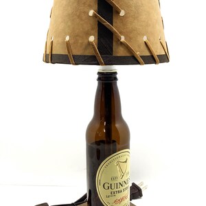 May include: A unique lamp crafted from a Guinness beer bottle, featuring a brown lampshade with decorative stitching. The lamp sits on a wooden base and includes a power cord and plug.