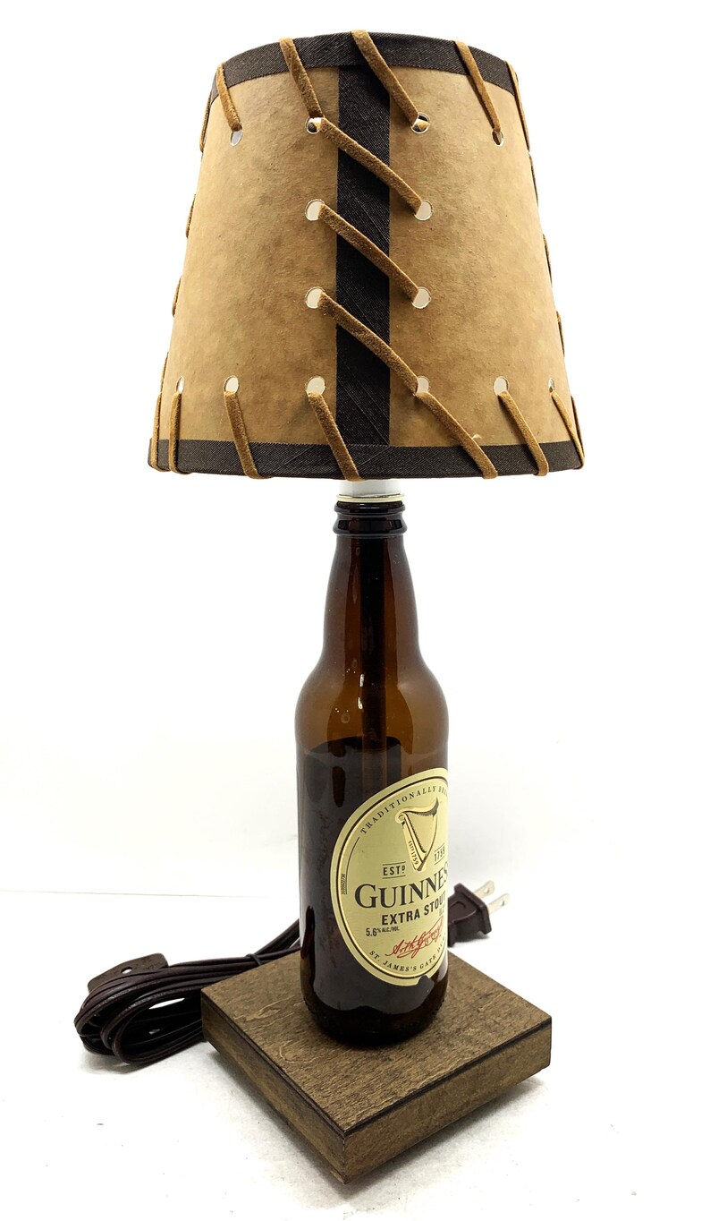 May include: A unique lamp crafted from a Guinness beer bottle, featuring a brown lampshade with decorative stitching. The lamp sits on a wooden base and includes a power cord and plug.