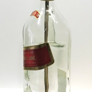 Puede incluir: Una l&aacute;mpara de botella de vidrio transparente con una pantalla dorada y una base de madera. La botella tiene una etiqueta que dice "Johnnie Walker Red Label Blended Scotch Whisky."