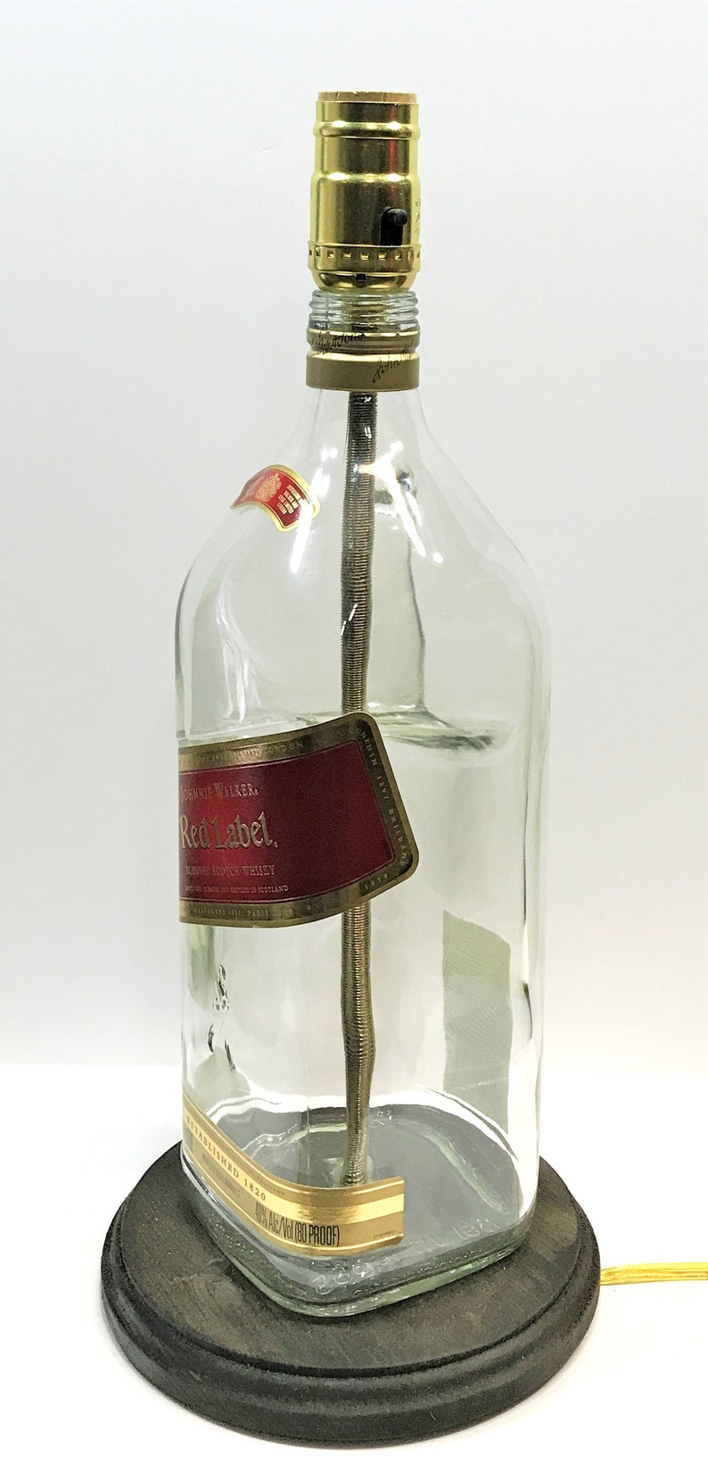 Puede incluir: Una l&aacute;mpara de botella de vidrio transparente con una pantalla dorada y una base de madera. La botella tiene una etiqueta que dice "Johnnie Walker Red Label Blended Scotch Whisky."