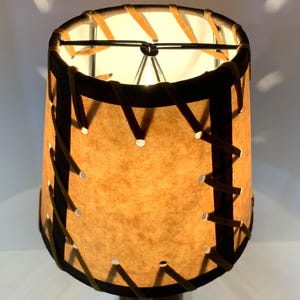 May include: A cylindrical lampshade with a tan, textured surface and dark brown trim. The shade is stitched with a lighter brown material. The lamp is lit from within, casting a warm glow. The lamp is a decorative home decor item.