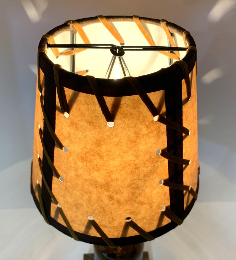 May include: A cylindrical lampshade with a tan, textured surface and dark brown trim. The shade is stitched with a lighter brown material. The lamp is lit from within, casting a warm glow. The lamp is a decorative home decor item.