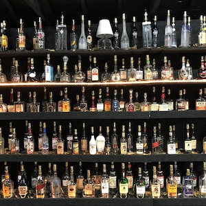 May include: A collection of liquor bottles displayed on black shelves. The bottles vary in shape, size, and color, with a variety of labels. The shelves are illuminated with a warm light, creating a bar or liquor store aesthetic.