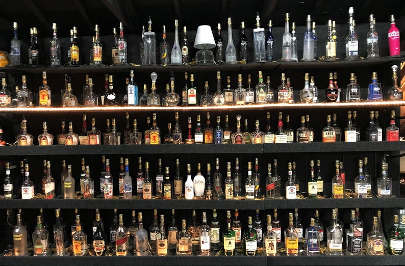 May include: A collection of liquor bottles displayed on black shelves. The bottles vary in shape, size, and color, with a variety of labels. The shelves are illuminated with a warm light, creating a bar or liquor store aesthetic.