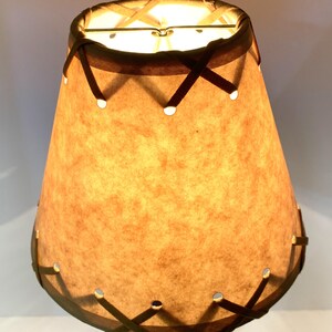 May include: A vintage-style lampshade with a warm, tan paper shade. The shade is cone-shaped with a dark brown trim and decorative cross-stitched accents. Small circular cutouts are evenly spaced around the bottom edge. The lamp is lit from within, casting a warm glow.