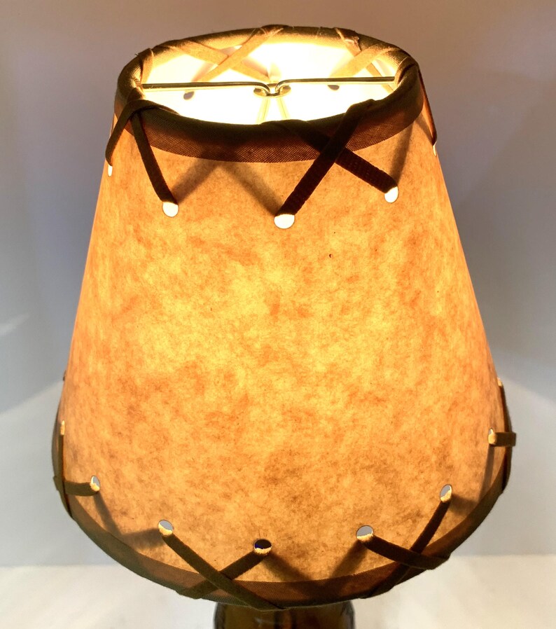 May include: A vintage-style lampshade with a warm, tan paper shade. The shade is cone-shaped with a dark brown trim and decorative cross-stitched accents. Small circular cutouts are evenly spaced around the bottom edge. The lamp is lit from within, casting a warm glow.