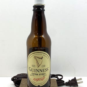 May include: A brown Guinness Extra Stout beer bottle with a white top, resting on a small wooden base. The bottle has a label with the Guinness logo and text. A brown electrical cord and plug are visible.