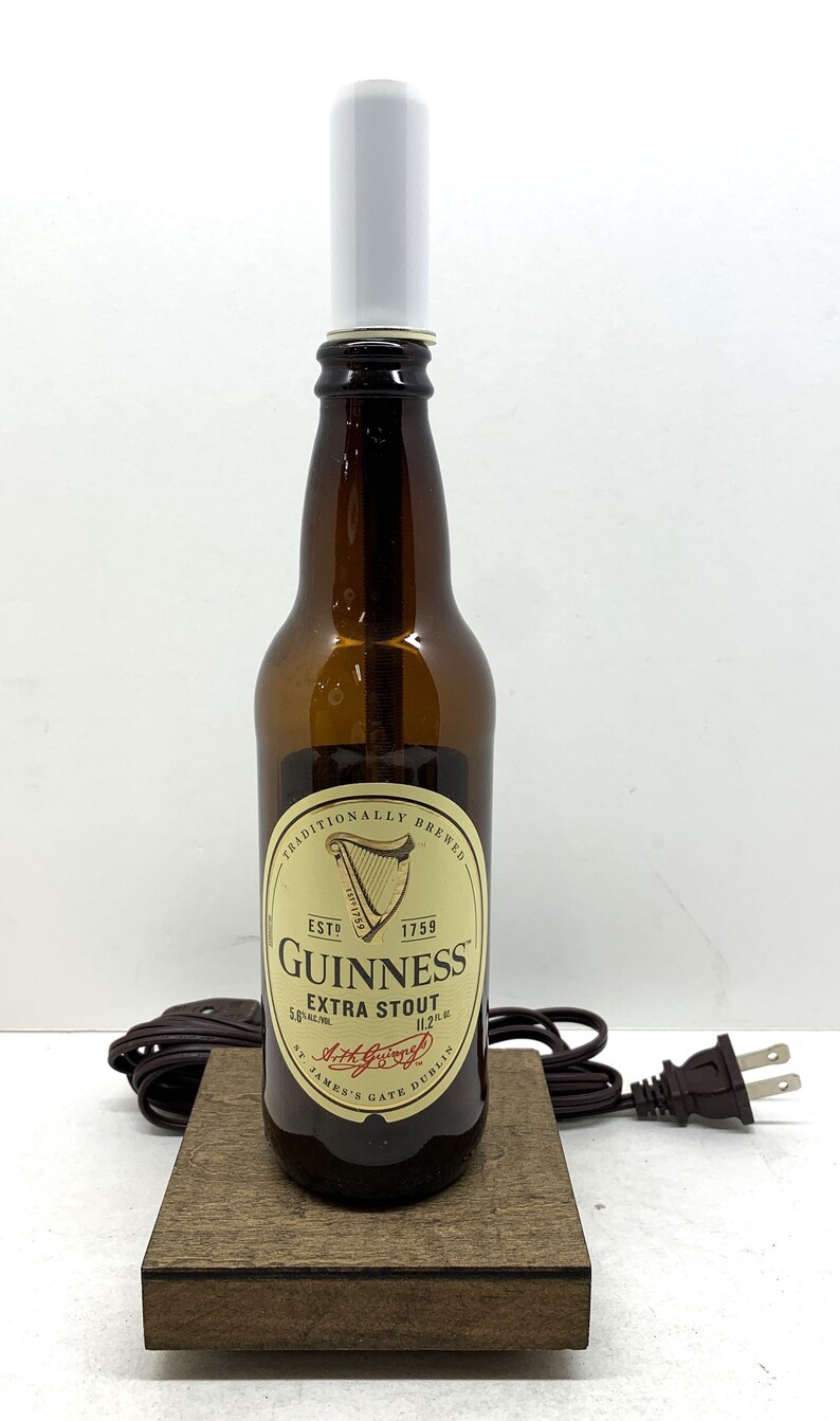 May include: A brown Guinness Extra Stout beer bottle with a white top, resting on a small wooden base. The bottle has a label with the Guinness logo and text. A brown electrical cord and plug are visible.