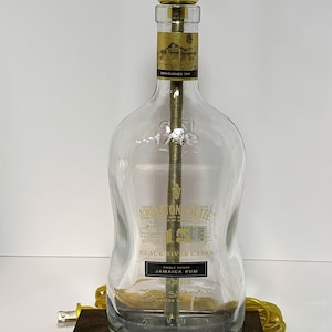 May include: A clear glass bottle repurposed as a lamp. The bottle has a gold label with text and a gold lamp fixture. The lamp sits on a dark brown wooden base and has a yellow cord.