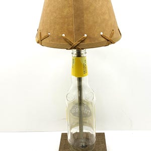 May include: A unique table lamp crafted from a repurposed glass bottle, featuring a tan lampshade with decorative stitching. The lamp sits on a wooden base and includes a power cord with a plug.