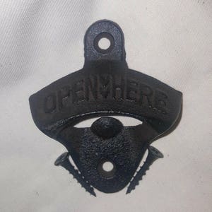 Puede incluir: Abrebotellas de hierro fundido negro con las palabras "Open Here" en relieve en la parte superior. El abrebotellas tiene dos tornillos en la parte posterior para montarlo.