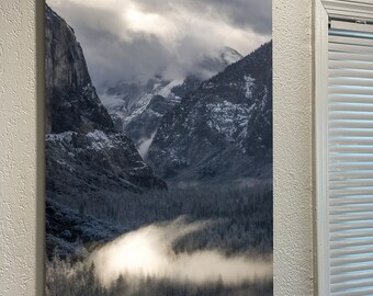 May include: A panoramic view of a snow-capped mountain range with a valley filled with fog and trees. The sky is cloudy and the sun is shining through the clouds.