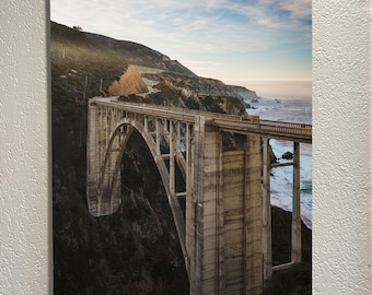 May include: A concrete bridge arches over a rocky coastline with a view of the ocean. The bridge is a popular tourist destination and offers stunning views of the Pacific Ocean.