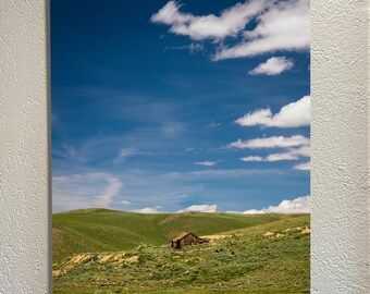 May include: A small wooden cabin sits on a grassy hillside under a bright blue sky with fluffy white clouds.