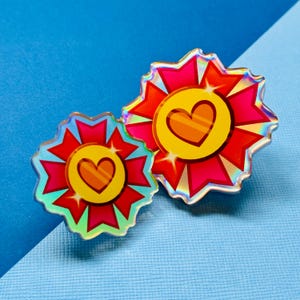 May include: Two iridescent holographic enamel pins shaped like red and yellow flowers with a heart in the center. The heart is orange with a yellow outline and a white stripe.