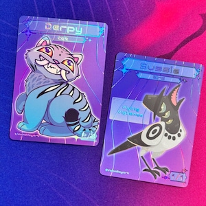 May include: Two holographic trading cards with cartoon animal illustrations. One card features a blue cat with the name "Derpy" and the other shows a black and white bird named "Sussie". The cards have a purple and blue background.