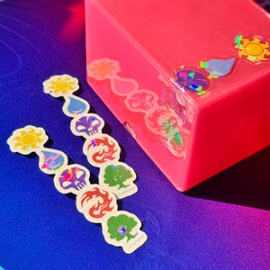 May include: A collection of colorful stickers featuring various symbols, including suns, water droplets, skulls, flames, and trees. The stickers are arranged on a blue surface next to a pink box. The stickers have a holographic effect.