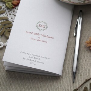 May include: A white notebook with a pink logo that says "SLG Good Little Notebooks by Some Little Good". The notebook features a watercolor print of Beth March by Bridget A. Deputy. Copyright 2021.