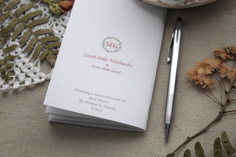 May include: A white notebook with a pink logo that says "SLG Good Little Notebooks by Some Little Good". The notebook features a watercolor print of Beth March by Bridget A. Deputy. Copyright 2021.