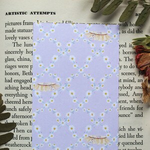May include: A purple bookmark with a white daisy pattern. The bookmark has a white lace trim and the text "215" is written on the lace.