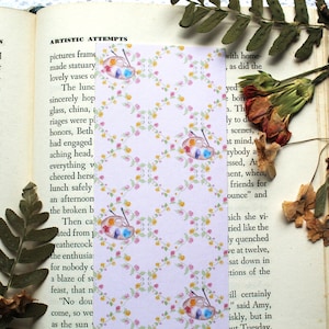 May include: A pink and white bookmark with a repeating pattern of small flowers and watercolor paint palettes. The bookmark is made of paper and has a slightly textured surface.
