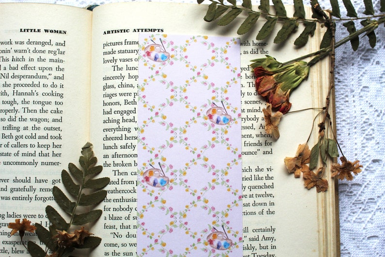 May include: A pink and white bookmark with a repeating pattern of small flowers and watercolor paint palettes. The bookmark is made of paper and has a slightly textured surface.