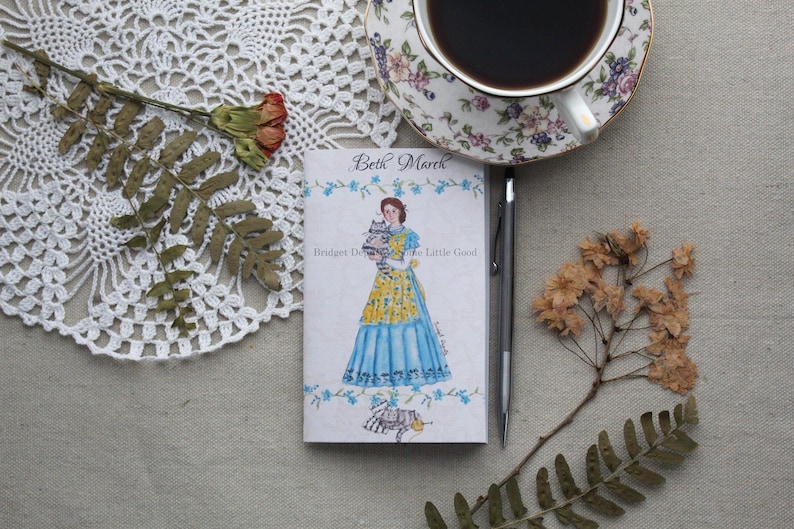May include: A small, white notebook with a watercolor illustration of a woman holding a cat. The notebook has the text "Beth March" and "Bridget Devey" on the front cover. The text "The Little Good" is also on the cover.
