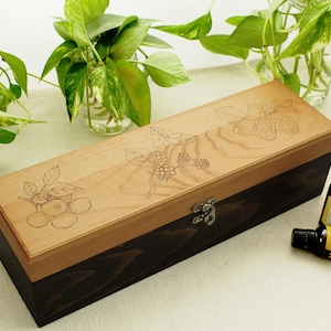May include: A wooden box with a light brown lid and dark brown base, featuring etched fruit designs. The box is rectangular with a metal clasp. Two small brown bottles with black caps are next to the box. Green plants in glass vases are in the background.