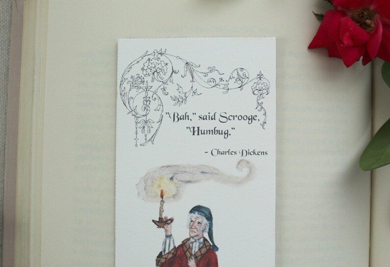 Pode incluir: Um marcador de p&aacute;gina com uma ilustra&ccedil;&atilde;o em preto e branco de um homem segurando uma vela. A cita&ccedil;&atilde;o "Bah," disse Scrooge. "Humbug." - Charles Dickens est&aacute; impressa no marcador de p&aacute;gina.