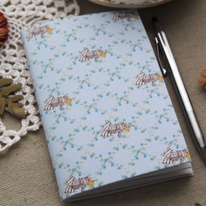 May include: A blue notebook with a floral pattern featuring small white flowers and a repeating image of a small animal with a yellow object in its mouth. A silver pen is lying next to the notebook.