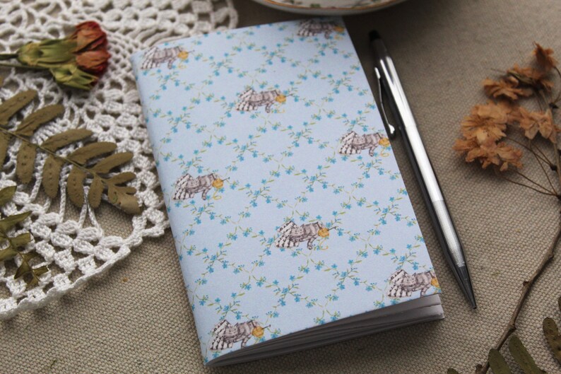 May include: A blue notebook with a floral pattern featuring small white flowers and a repeating image of a small animal with a yellow object in its mouth. A silver pen is lying next to the notebook.