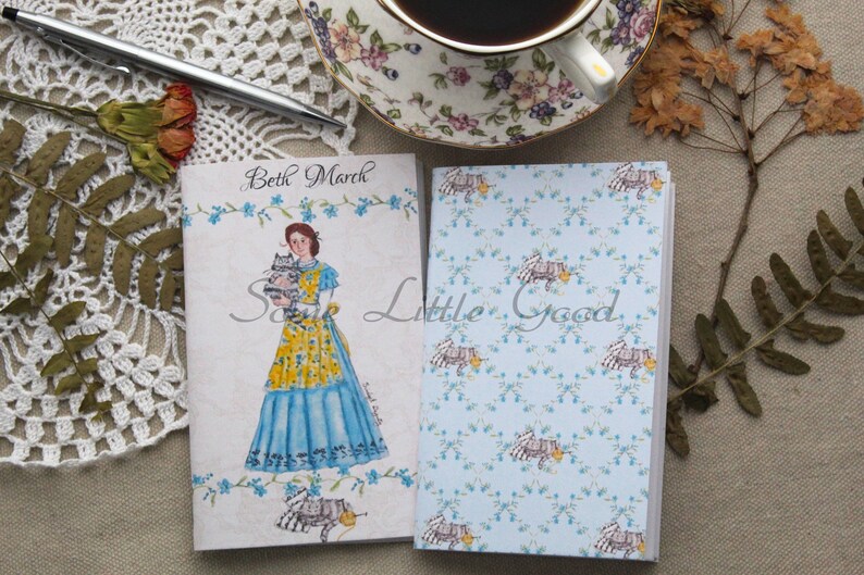 May include: Two small notebooks with a blue and white floral pattern. The front cover of one notebook features a watercolor illustration of a woman in a blue dress holding two kittens. The text "Beth March" is printed on the front cover of the notebook.