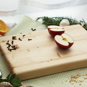 May include: A light-colored wooden cutting board with a rectangular shape. Halved red apples, orange slices, and cloves are arranged on the board. A small knife and sprigs of rosemary and other herbs are also present.