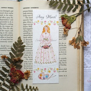 May include: A watercolor bookmark featuring a girl in a pink dress holding a bouquet of flowers. The bookmark has the text "Amy March" and "Some Little Good" on it. The background is a light blue with a watercolor painting of a palette and paintbrushes.